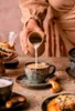A person is pouring milk into a coffee cup, with pastries and coffee brewing equipment in the background. Podgląd zdjęcia produktu