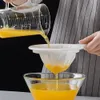 A person is pouring a yellow liquid from a glass measuring cup through a white mesh sieve into a glass bowl. Produktbild-Vorschau
