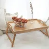 A wooden tray table with a basket of bread and a vase with eucalyptus on a fluffy rug, near some pillows. Produktbild-Vorschau
