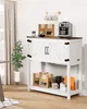 A white farmhouse-style cabinet with a wooden top, used as a coffee bar in a dining or living area. It features two cabinet doors with barn-style X-detailing and black hardware. The top surface holds a coffee maker, coffee bags, and a small plant. The open Produktbild-Vorschau