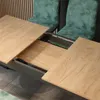 A close-up view of an extendable dining table with two green chairs in the background. Produktbild-Vorschau