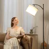 A woman is sitting on a couch reading a book next to a floor lamp. Voir les images du produit