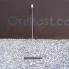 A metal tool with a flat base and a long handle stands upright on a bed of gravel in front of a rusty metal wall with a faded logo. Podgląd zdjęcia produktu