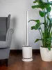 A white tower fan is placed on a wooden floor next to a grey armchair and a large potted plant. Podgląd zdjęcia produktu