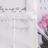 A close-up view of a white fabric with handwritten text and a clear plastic tube resting on it, with pink roses in the background. Produktbild-Vorschau