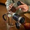 A person is preparing coffee using a French press and an electric coffee grinder. Produktbild-Vorschau