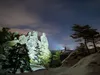 A person stands on a rocky outcrop at night, shining a flashlight into the distance. The scene is set against a starry sky with clouds and illuminated rock formations. Produktbild-Vorschau