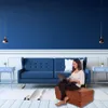 A woman is working on her laptop while sitting on a folding ottoman in a living room with a blue couch. Produktbild-Vorschau