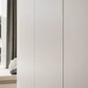 A minimalist interior scene with a large white wardrobe dominating the frame. To the left, a glimpse of a window with grey curtains and a cushioned window seat with a patterned throw. Produktbild-Vorschau