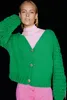 A woman wearing a green cardigan and pink turtleneck and pants against a black background. Produktbild-Vorschau