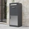 A dark gray parcel mailbox with silver accents is shown outdoors, standing on a tiled patio next to a stone wall and a window. Produktbild-Vorschau