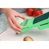 A person's hands are using a green vegetable slicer on a white table. Two red tomatoes and a potato are visible in the background and foreground. Produktbild-Vorschau