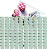 A large collection of small glass jars with green and white checkered lids, arranged in rows. In the background, there are pink flowers, a red and white checkered cloth, and a small packet with German text. Náhled obrázku produktu