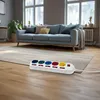 A power strip with multiple sockets is placed on a wooden floor in front of a sofa and a rug in a living room. Produktbild-Vorschau