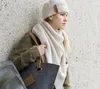 A young woman wearing a beige knit hat and scarf, leaning against a textured wall, holding a gray felt tote bag. Produktbild-Vorschau
