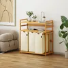 A wooden shelf unit with three beige laundry bags and decorative items on the shelves, next to a sofa and a large plant. Anteprima dell’immagine prodotto