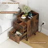 A rustic brown nightstand with two drawers is placed next to a bed. The top surface displays a cup of coffee, a vase of flowers, and a wireless charging station with a phone. Podgląd zdjęcia produktu