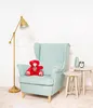 A red teddy bear sits on a light blue armchair next to a floor lamp and two small side tables. Náhľad obrázku produktu
