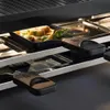 Close-up of a raclette grill with food in the upper tray and handles visible in the foreground and midground. Náhľad obrázku produktu