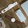 A white power strip with a red switch is plugged into an outlet, charging a phone and a laptop. A watch is also on the wooden table. Voir les images du produit