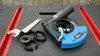 A set of angle grinder accessories, including a dust extraction shroud, a wrench, and several adapter rings, laid out on a black and red workbench. Náhled obrázku produktu
