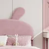 A close-up view of a pink upholstered headboard with bunny ears, a decorative pillow, and a stuffed bunny toy. Náhled obrázku produktu