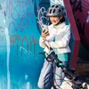 A young person wearing a helmet and holding a phone, standing next to a bicycle in front of a graffiti-covered wall. Produktbild-Vorschau