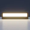 A long, rectangular LED wall light fixture is mounted on a wall, emitting a bright, warm white light downwards onto a surface. Produktbild-Vorschau