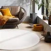 A cozy living room scene featuring a round rug, a comfortable armchair with cushions and a throw, and decorative items near a window.