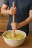 A person is using a wooden masher to mash shredded ingredients in a large white bowl. Produktbild-Vorschau