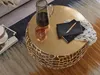 A round gold coffee table with a decorative base, placed on a rug in a living room setting. A pitcher, a glass, and a notebook with a pen are on the table. Produktbild-Vorschau