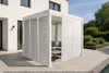 A modern white pergola with louvered walls and a seating area inside, set on a tiled patio next to a house and garden. Náhľad obrázku produktu