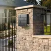 A modern mailbox attached to a stone pillar in front of a house and fence. Voir les images du produit