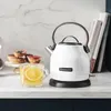 A white KitchenAid electric kettle sits on a marble countertop next to a glass mug of tea with lemon slices and a small plate of tarts. Produktbild-Vorschau