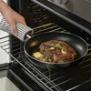 A hand holding a striped towel is placing a frying pan with a steak into an oven. Produktbild-Vorschau