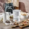A coffee grinder, a foldable travel kettle, coffee beans, and dried fruit are arranged on a textured surface, with a drawstring bag in the background. Produktbild-Vorschau