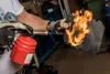 A person is using a blowtorch to heat a piece of black material, possibly for shaping or sealing. Produktbild-Vorschau