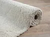 A close-up view of a rolled-up shaggy rug with a cream-colored, high-pile texture, resting on a wooden floor. Produktbild-Vorschau