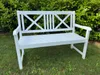 A white wooden garden bench sits on a grassy lawn in front of a green hedge. Produktbild-Vorschau