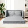 A grey sofa bed is laid out flat on a rug in a well-lit room, with a large potted plant on the left and a bookshelf on the right. Produktbild-Vorschau