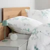 A close-up view of a bed with white bedding featuring a green floral pattern, a wooden headboard, and a bedside table with a lamp and vase. Produktbild-Vorschau