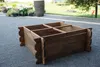 A wooden planter box with multiple compartments sits on a paved patio in front of a garden bed and a large stone pot. Náhled obrázku produktu