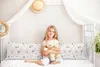 A young girl sits on a bed holding a teddy bear, with a large pillow in front of her. Produktbild-Vorschau