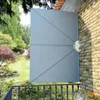 A gray folding patio side awning is attached to a brick wall, overlooking a lush garden and balcony railing. Produktbild-Vorschau