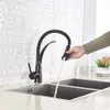 A person's hand is shown using a black kitchen faucet with a pull-down sprayer, with water running into a double stainless steel sink. The background features a white brick-patterned wall. Produktbild-Vorschau