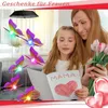 A mother and daughter are looking at a handmade card with the word 'MAMA' and a heart drawn on it. The daughter is holding the card, and the mother is holding a bouquet of tulips. Butterfly-shaped wind chimes with colorful LED lights are hanging in the bac Produktbild-Vorschau