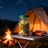 Transparentes Glas-Mason-Glas mit grünem Deckel, gefüllt mit geschichtetem Salat, auf einem Campingtisch neben gegrillten Würstchen, mit Lagerfeuer und Zelt im Hintergrund unter sternenklarem Nachthimmel Podgląd zdjęcia produktu