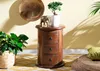 A wooden nightstand with drawers, decorated with a bonsai tree, an elephant figurine, and a plant in a basket, set against a yellow wall with shadows. Voir les images du produit