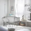 A macrame hanging chair with cushions is set up in a bright, airy room near a window with blinds. Voir les images du produit