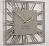 A modern gray square wall clock with Roman numerals and gold hands, mounted on a white wall. Náhled obrázku produktu